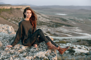 Serene woman in black sweater and brown boots sitting on rock at mountain summit