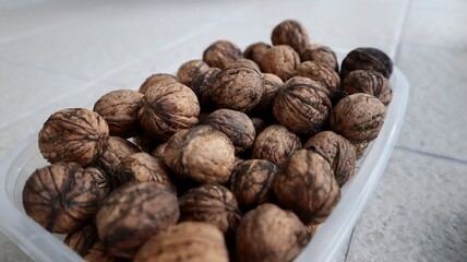 A close-up of a plastic container filled with whole walnuts. The walnuts have dark lines and strings (known as peduncles or fibers) on their shells, which are remnants from the husks.