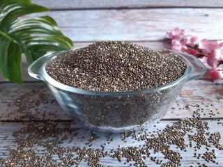 Clear Bowl of Black and White Chia Seeds on Wood, with Glossy Texture, Scattered Seeds, and Pink Flowers Adding a Decorative Touch in the Background.