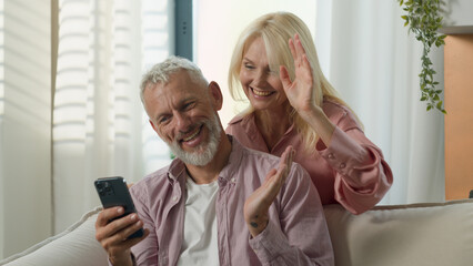 Cheerful Caucasian old senior elderly couple family talking mobile phone gadget man male woman female online video call wife husband talk blogging vlog together waving hands greeting inside home room
