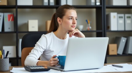 A young woman at her desk gazes thoughtfully into the distance, taking a break from work on her laptop, surrounded by books and office supplies.