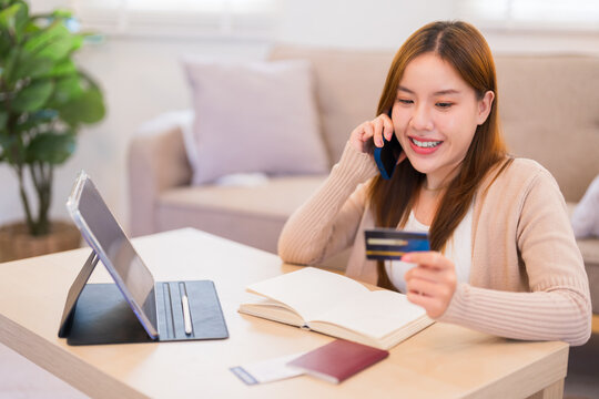 A young woman is sitting at a table, talking on the phone while holding a credit card, likely making a travel purchase, with a tablet, passport, and notebook on the table - Powered by Adobe