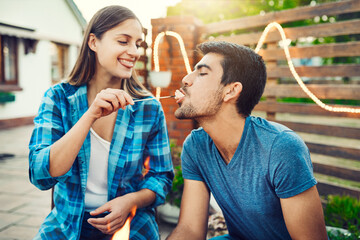 Feeding, marshmallow and playful with couple at party together for barbecue, celebration or love. Biting, eating or flirting with happy man and woman outdoor for bonding, relationship or romance