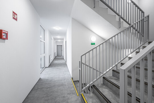 Modern hallway with stairs and metal railings in a bright building, featuring geometric lines and a long corridor leading to multiple doors.