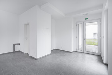 Spacious, modern, and minimalistic room with large windows, white walls, and gray tiled floors, featuring clean lines and a bright, open feel, leading to an outdoor area.