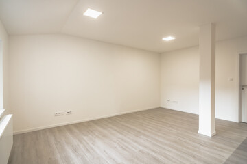 A spacious, empty room with light-colored walls and parquet flooring, featuring modern ceiling lights and a single supporting pillar in the center.