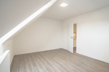 Bright attic room with angled ceiling, a single window, and an open door leading to a kitchen. The space is empty, with light-colored parquet flooring and a modern design.