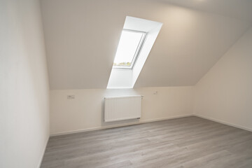 A minimalistic attic room with a single window, slanted roof, and light-colored parquet flooring....