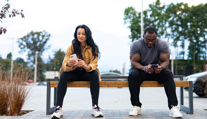 Fototapeta premium Two people using smartphones on a bench in a public area. A man and a woman sit on a bench, both focused on their smartphones in a relaxed setting.
