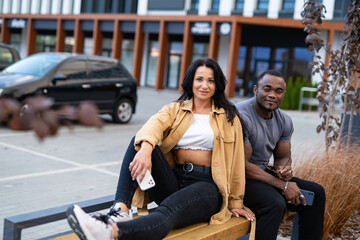 Fototapeta premium Couple relaxing on a bench outside a modern building in daylight. A couple sits on a bench, enjoying time together outside a contemporary structure.