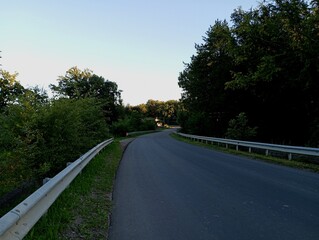 Asphalt road with metal bumpers along the roadside. Trees and bushes along the road. A small bend in the road.
