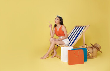 Happy Asian sexy woman dressed in summerwear with shopping bag sitting on beach chair pointing...