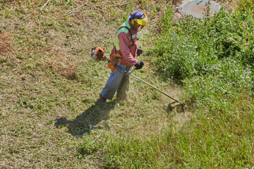 Summer heat doesn't stop the gardener in overalls and a helmet from working with a manual lawnmower on the overgrown lawn.