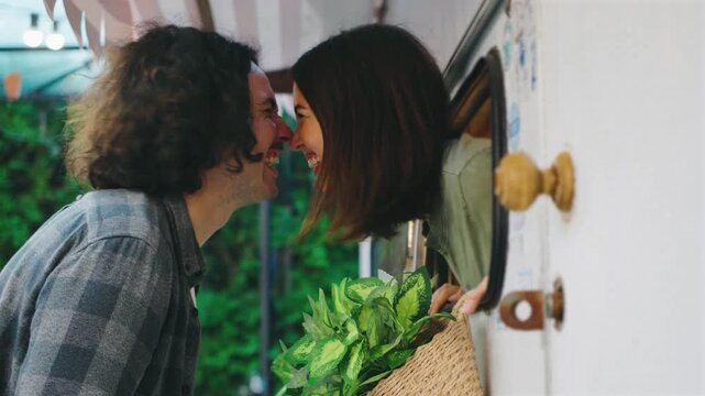 A girl looks out the camper window, playfully rubbing noses with her boyfriend outside. The scene is set amidst lush greenery, creating a cozy, loving atmosphere.