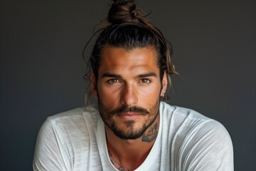 Obraz premium Handsome man with Top Knot hair style in casual white shirt gazing intensely at the camera against dark background. Concepts include self-confidence, attractiveness, and individuality.