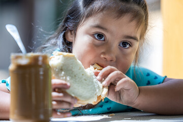 A young girl is eating a sandwich with a jar of peanut butter next to her. The scene is casual and...