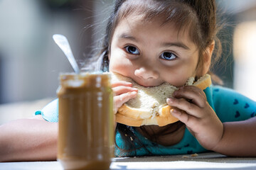 A young girl is eating a sandwich with a jar of peanut butter next to her. The scene is casual and relaxed, with the girl enjoying her snack