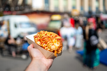 A hand holding a hot dog with sauce in a paper tray, set against a blurred urban street background. The image captures the essence of street food culture with a focus on the delicious snack.