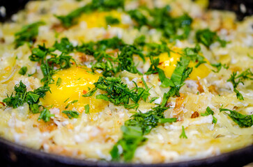 Fried potatoes in a cast iron frying pan with three broken chicken eggs. Three whole yolks are sprinkled with finely chopped arugula and dill.