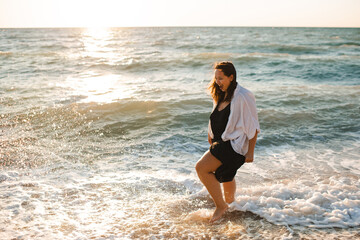 Smiling beautiful overweight woman 30 - 35 year old wearing black dress walking at beach over sea close up. Body positive concept.