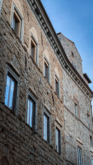 Sunlit medieval stone facade with ornate windows and cornices. Textured walls reveal centuries of history. Blue sky frames timeless Italian architecture.