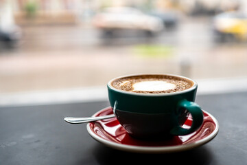 Close-up of a cappuccino cup with heart-shaped latte art on a saucer with a spoon, set against a blurred street view background. Perfect for themes of coffee, relaxation, and urban lifestyle.