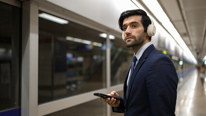 Project manager wearing headphone at train station while holding mobile phone for choosing song. Smart business man listening relaxing music while waiting for train with blurred background. Exultant.