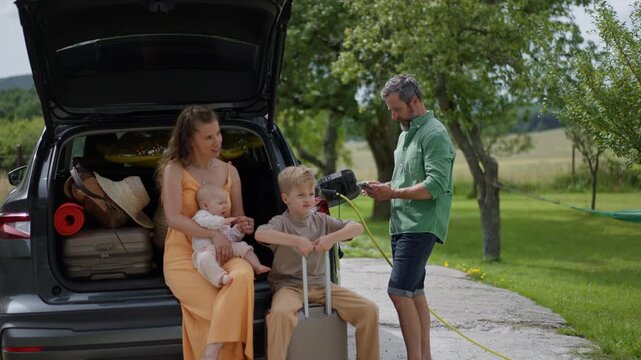 Family loading luggage into trunk of electric car with charger in charging port. Travelling on family road trip or vacation by electric vehicle.