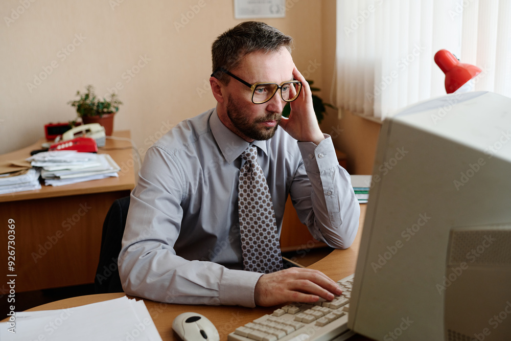 Poster portrait of a caucasian man sitting at desk, using a computer in an office environment, wearing glas