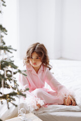 A young girl is sitting on a bed in front of a Christmas tree. She is wearing a pink pajama set and she is looking at the camera. Concept of warmth and coziness