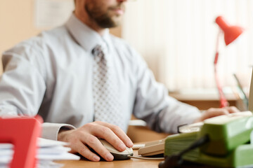 Person working at office desk, using a computer and vintage telephone, surrounded by office...