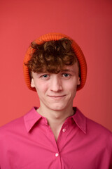 Portrait of young Caucasian man smiling and wearing a bright orange beanie and pink shirt against a solid red background, creating a colorful and cheerful atmosphere