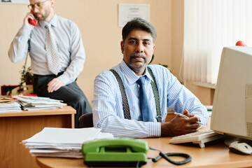 Businessman sitting at office desk in front of computer, another colleague talking on phone in background. Office filled with papers, ring binder, and necessary work devices