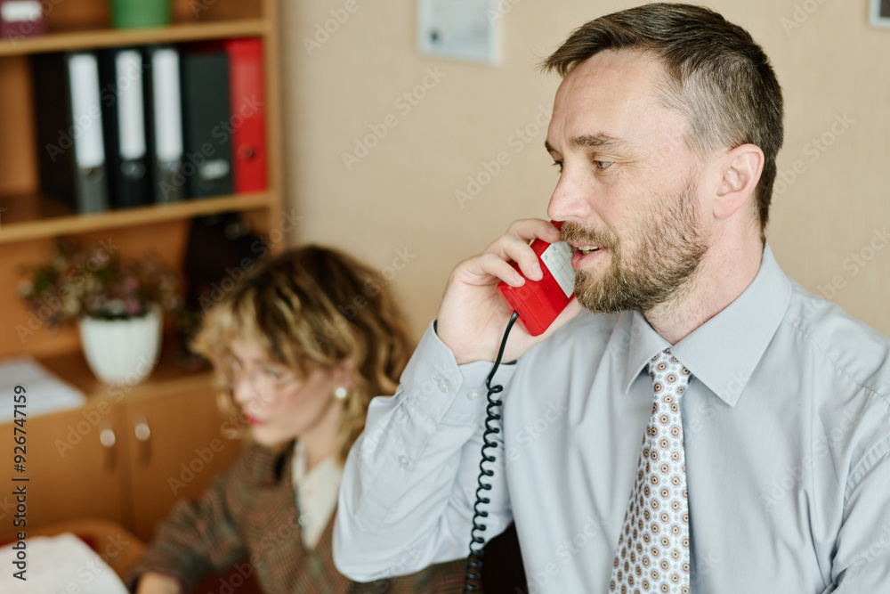 Poster Man in formal attire discussing projects over red wired landline phone while a woman in background focuses on her computer work on desk behind him