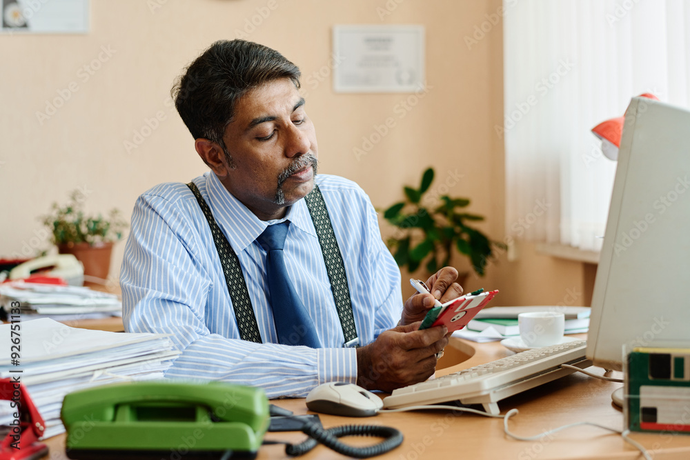 Poster indian man sitting at desk in an office environment, using a smartphone with intense concentration, 