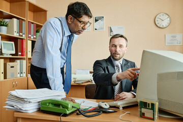 Two business professionals collaborating on work tasks in office setting, with documents and computer visible on desk. One man pointing at screen, other listening intently