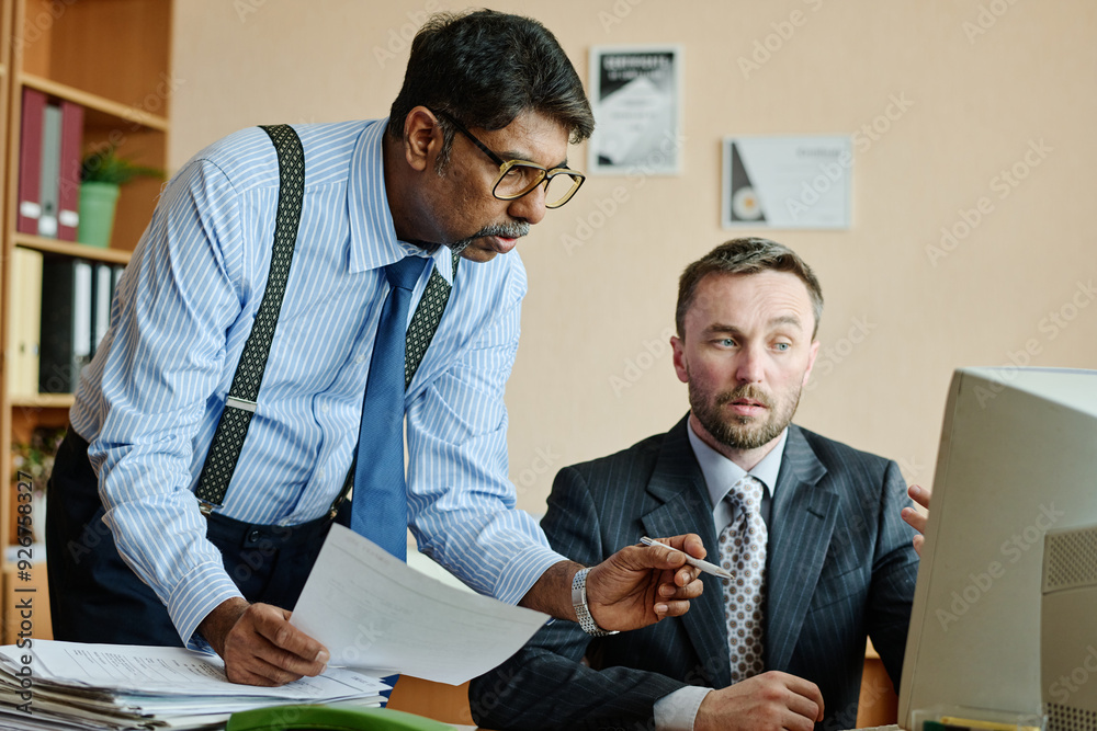 Poster Two business professionals are discussing strategies while looking at documents and computer. Office environment with shelves and office supplies in background