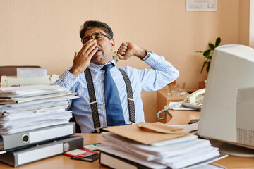 Businessman wearing striped shirt and suspenders yawning and stretching at desk with stacks of paperwork and office equipment around him in corporate environment