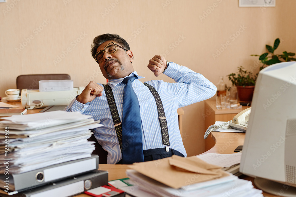 Poster businessman stretching arms at desk in an office filled with papers and documents, taking a break fr