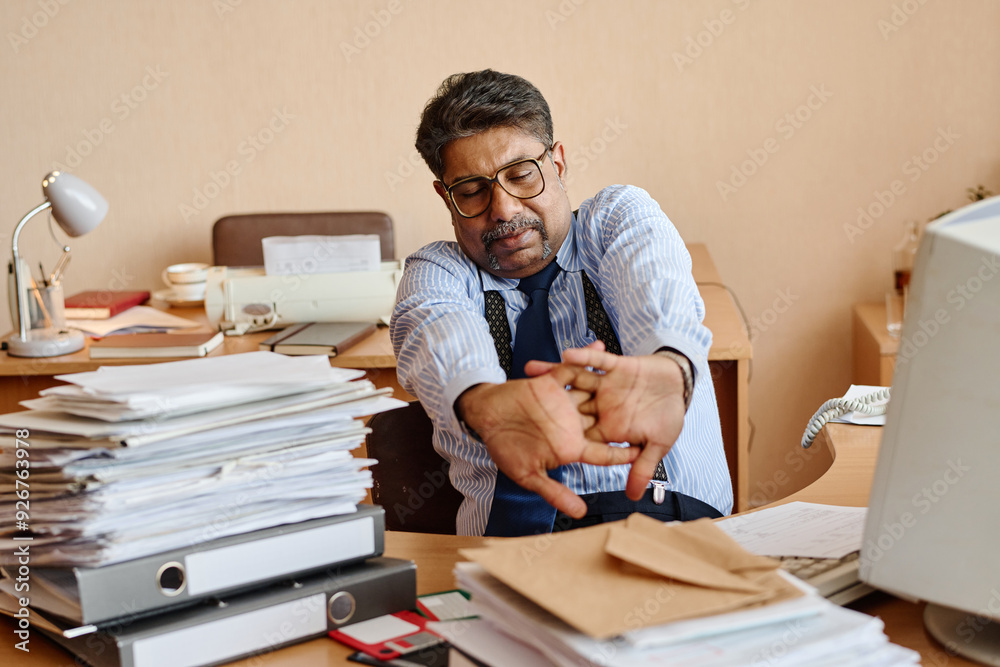 Poster stressed office worker stretching arms while sitting at cluttered desk with documents and files. off