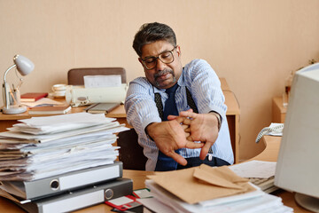 Stressed office worker stretching arms while sitting at cluttered desk with documents and files. Office environment showing piles of paperwork, computer, and traditional lamp