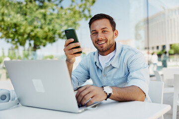 Smiling young man using smartphone and laptop outdoors at a cafe, enjoying technology and connectivity