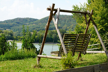 A wooden swing set in a picturesque Austrian rural area, nestled in green grass with a clear sky, trees, and a serene river nearby