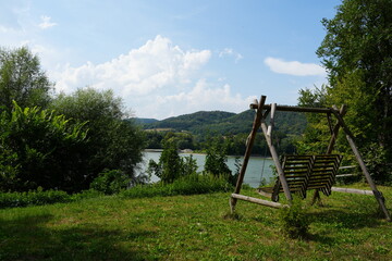 Fototapeta premium A wooden swing in a scenic Austrian countryside, surrounded by vibrant green grass, a clear blue sky, trees, and a calm river