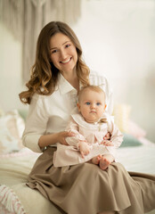 Beautiful young mother sits on the bed and holds her infant daughter on lap in delicate pastel colors interior. Maternal love and child care