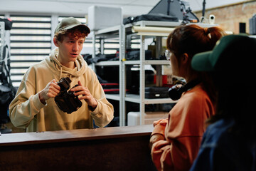 Young man wearing a cap and holding a camera, conversing with two other people in a well-equipped studio with shelves in background