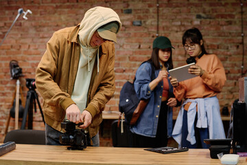Man adjusting camera on desk while two team members in background are discussing project using tablet in modern photography studio with brick wall interior