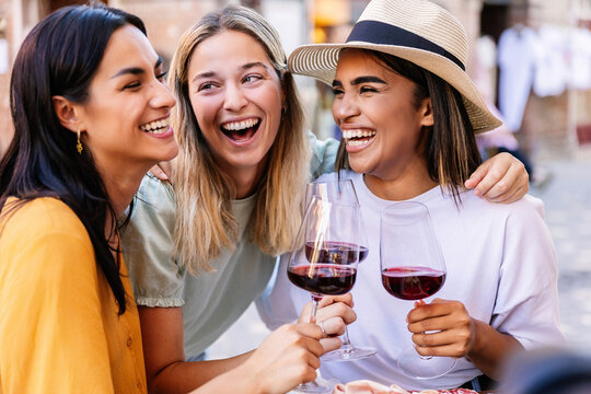 Three young multiracial women having fun enjoying aperitif with red wine in a restaurant. Female friendship and vacation lifestyle concept. - Powered by Adobe