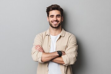 Portrait of a smiling young man in a beige shirt, standing with arms crossed, looking casually at the camera against a gray background.