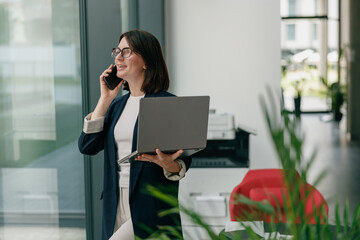 A businesswoman is talking on the phone while holding a laptop in a modern office